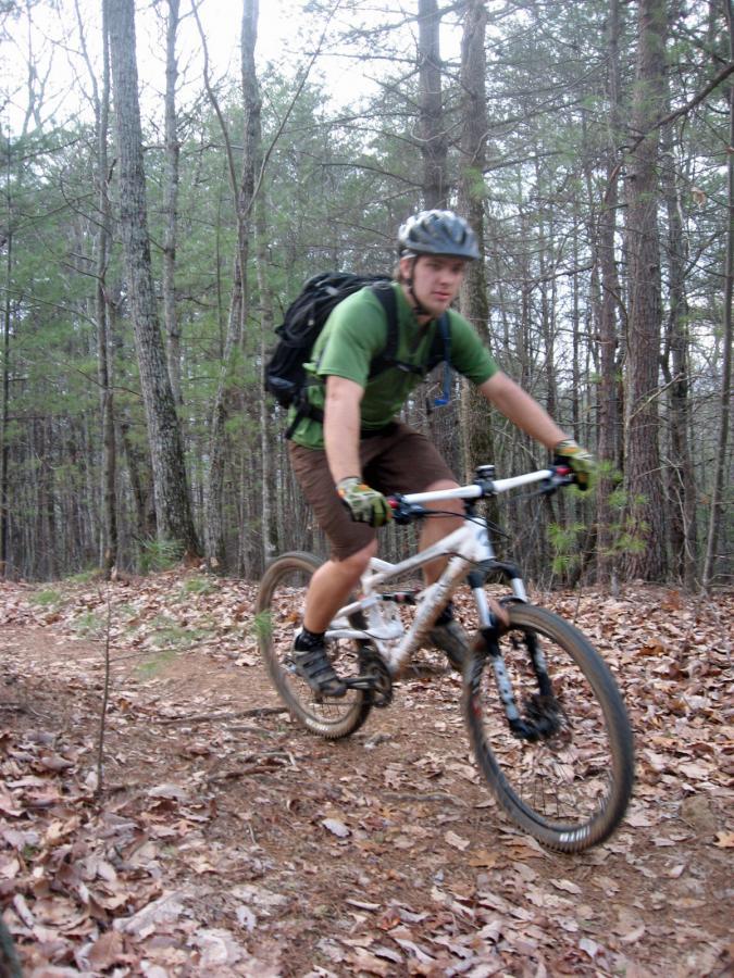 A person riding a mountain bike on a dirt trail surrounded by trees, wearing a helmet and a green shirt, with a backpack on their back. The ground is covered with leaves, and the cyclist is in motion, navigating the trail. Black Branch mountain bike trail.