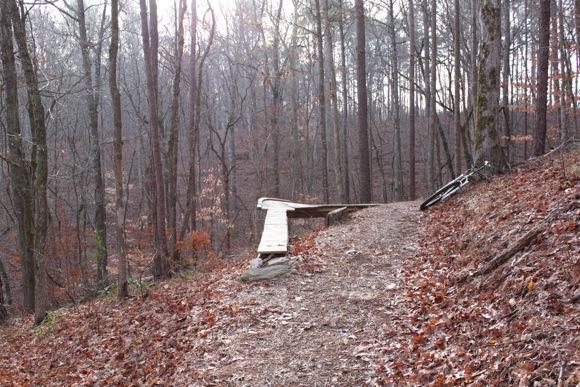 A wooded trail with a wooden ramp on the left side, surrounded by bare trees and autumn leaves scattered on the ground. A bicycle is leaning against the hillside near the ramp. The scene is softly illuminated by natural light filtering through the trees. Sylaward mountain bike trail.