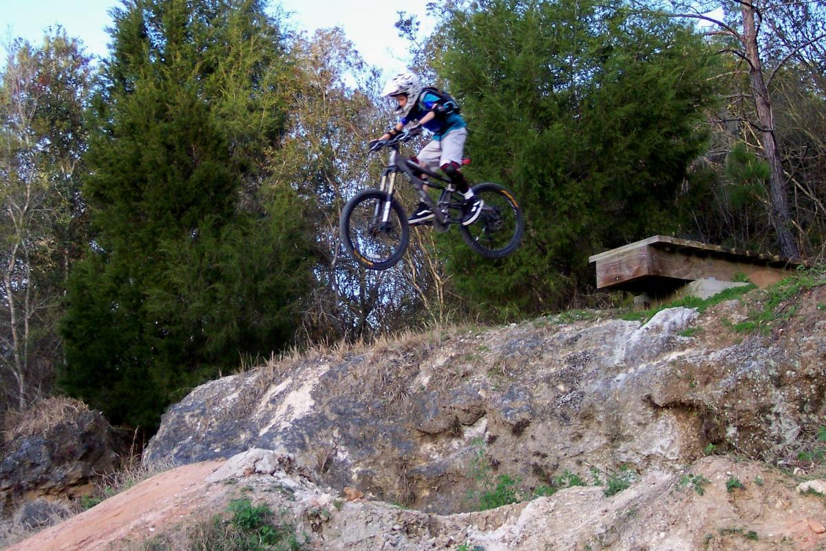 A young cyclist wearing a helmet and protective gear performs a jump on a mountain bike from a dirt ramp, with a backdrop of trees and rocky terrain. Santos mountain bike trail.
