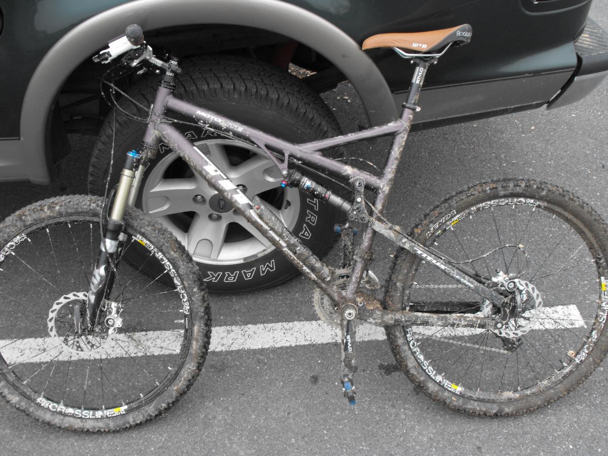 A muddy mountain bike leaning against a tire, parked on a pavement. The bike features thick tires and a specialized suspension system, showing signs of recent off-road use. 3rd Battle Of Winchester Trail mountain bike trail.