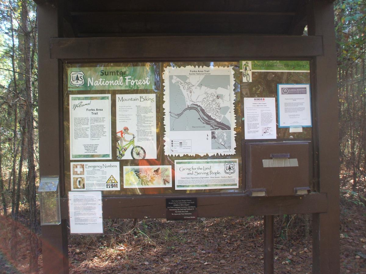 Signpost displaying information about the Forks Area Trail in Sumter National Forest. The board includes trail maps, guidelines for mountain biking, emergency numbers, and various notices. It is surrounded by trees and located in a natural setting. Forks Area Trail System (FATS) mountain bike trail.
