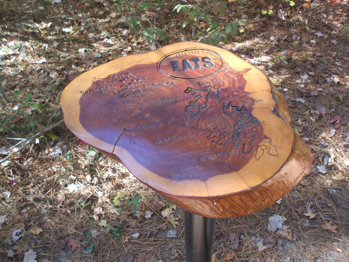 A wooden map plaque of the EATS trail system, featuring carved trail names and geographical details, mounted on a black metal pedestal, surrounded by natural foliage and leaves. Forks Area Trail System (FATS) mountain bike trail.