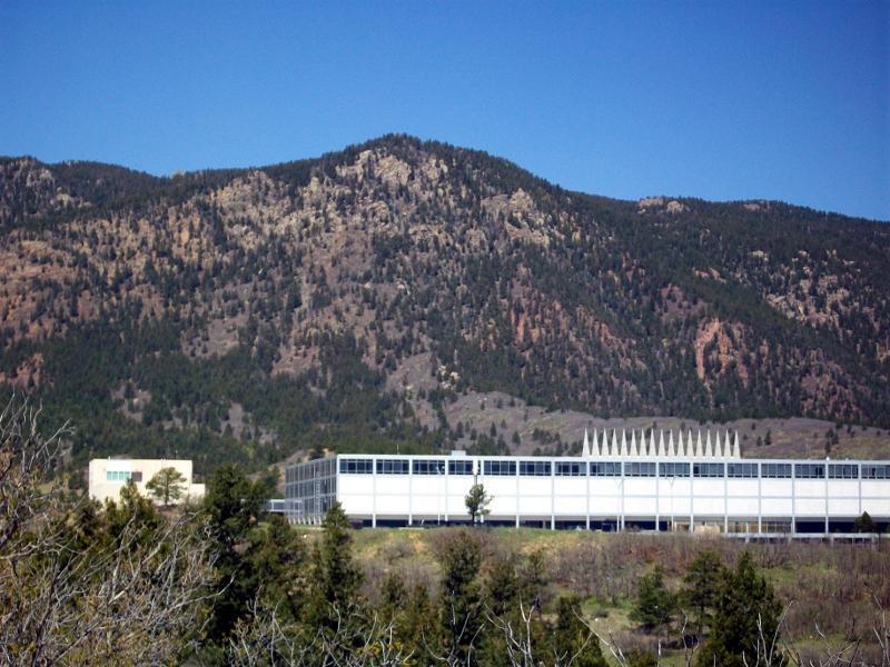 A panoramic view featuring a large, modern building set against a backdrop of rugged mountains and clear blue skies. In the foreground, various trees and shrubs are visible, adding greenery to the landscape. The building is characterized by rows of windows and a distinctive architectural design. Falcon Trail mountain bike trail.