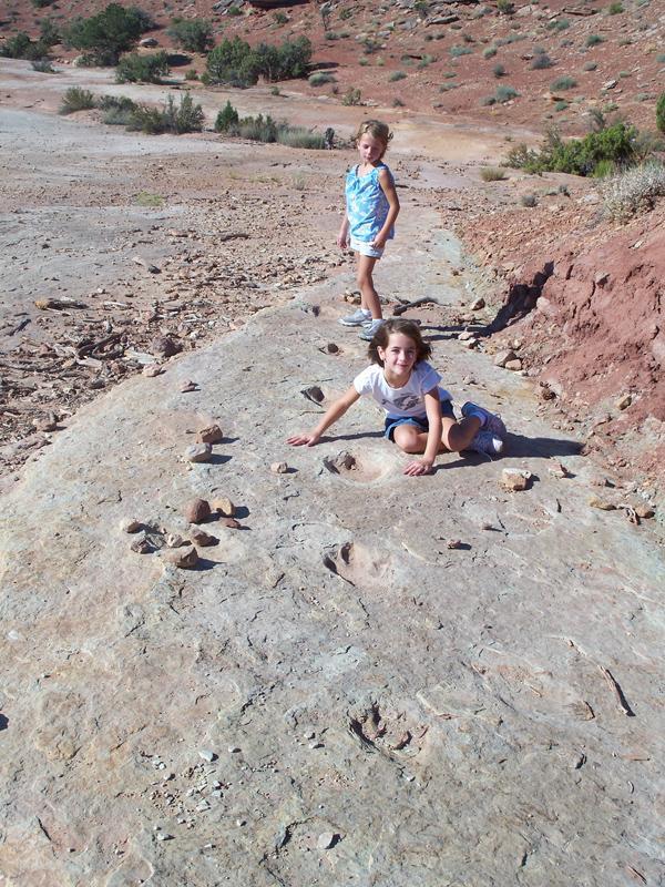 Two children exploring a rocky landscape. One child is crouched down on a flat rock surface, examining what appears to be fossilized footprints, while the other stands nearby. The background features a dry, mountainous area with sparse vegetation and red rock formations. Klondike Bluffs mountain bike trail.