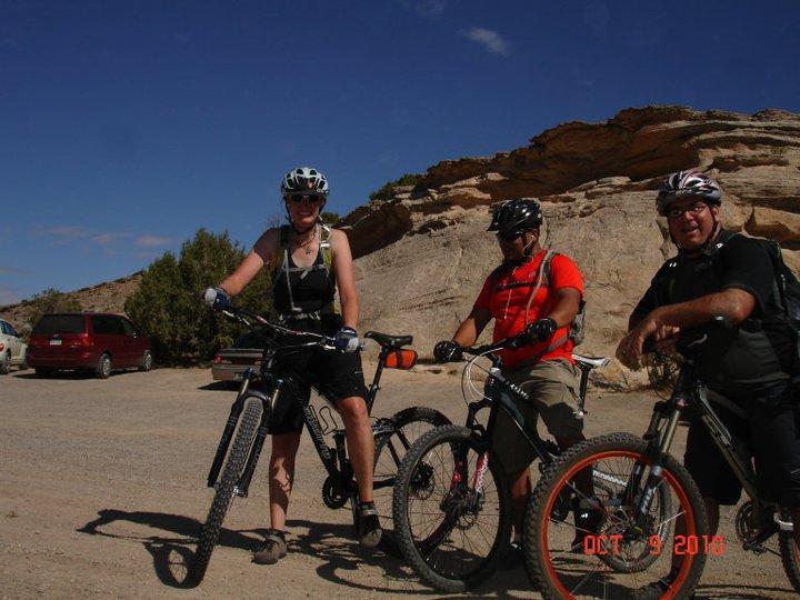 Three mountain bikers pose with their bicycles in a rocky outdoor setting. The first cyclist, wearing a helmet and sports gear, stands beside a black mountain bike. The second cyclist, in a red shirt, is holding a bike, and the third, wearing glasses and a dark shirt, is smiling next to an orange-accented bike. In the background, there's a clear blue sky and rugged rock formations. A parked vehicle is visible in the distance. Rustlers Loop mountain bike trail.
