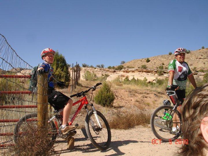 Two mountain bikers resting near a fence in a desert landscape. One rider leans against a post while the other stands beside their bicycle. The scene features sparse vegetation and rocky terrain under a clear blue sky. The first cyclist wears a blue shirt and red helmet, while the second is dressed in a green and white jersey. Rustlers Loop mountain bike trail.