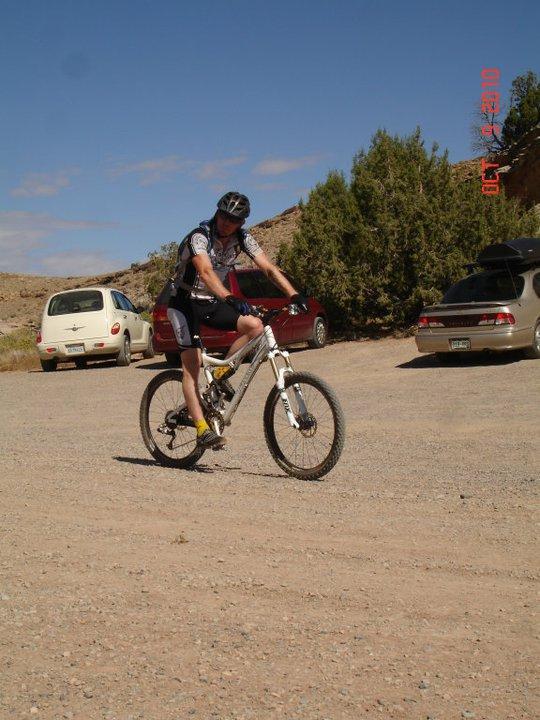 A person in cycling gear riding a mountain bike on a gravel surface, with parked cars and trees in the background under a clear blue sky. Rustlers Loop mountain bike trail.