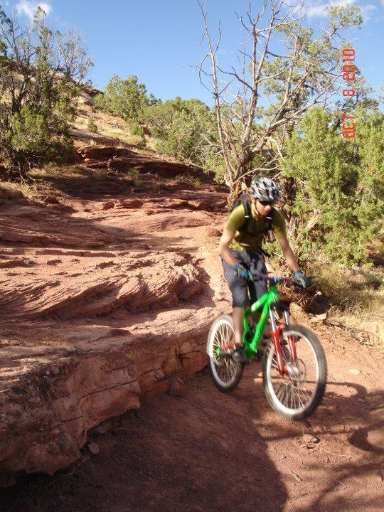 A mountain biker in a green and white bike jumps over a rocky terrain with trees in the background, against a clear blue sky. The rider is wearing a helmet and a backpack, navigating a rugged trail on a sunny day. Mary's Loop / Horsethief Bench mountain bike trail.