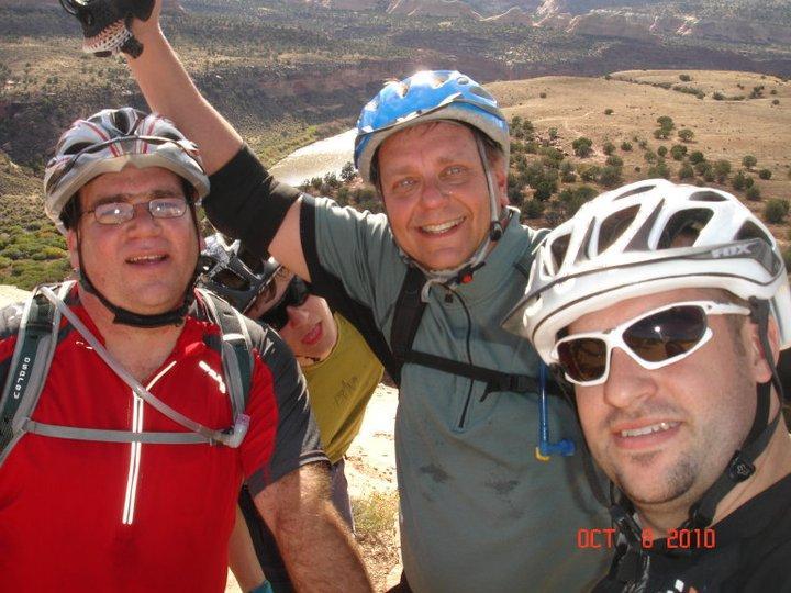 A group of four men smiling and posing for a selfie while mountain biking, wearing helmets and cycling gear. The background features a scenic landscape with hills and a river. The photo captures a moment of camaraderie outdoors. Mary's Loop / Horsethief Bench mountain bike trail.
