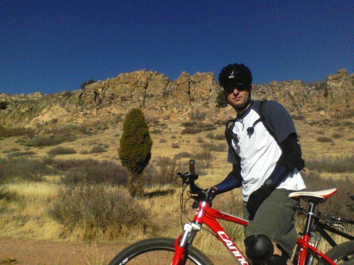 A person wearing a helmet and biking gear stands next to a red mountain bike on a dirt path. The background features rocky formations and sparse vegetation under a clear blue sky. Red Rock Canyon mountain bike trail.