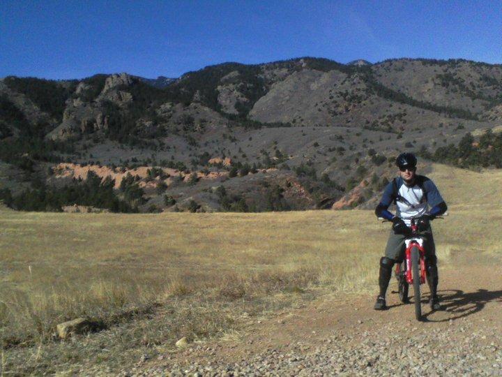 A person in cycling gear is riding a red mountain bike on a dirt trail surrounded by a vast landscape of grassy hills and rocky mountains under a clear blue sky. Red Rock Canyon mountain bike trail.