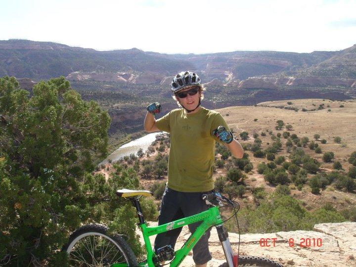 A person in a green mountain bike stands on a rock ledge, smiling and giving a thumbs-up. They are wearing a helmet, sunglasses, and biking gloves. In the background, a scenic landscape features rolling hills and a river winding through the valley below, under a clear blue sky. The date in the corner indicates October 8, 2010. Mary's Loop / Horsethief Bench mountain bike trail.