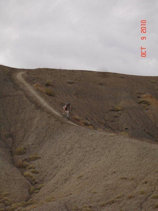 A mountain biker rides down a steep, winding dirt trail on a hilly terrain, with patches of vegetation scattered across a brown and gray landscape under a cloudy sky. Zippety Do Dah mountain bike trail.