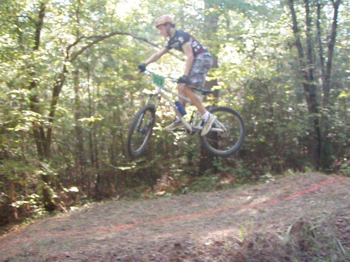 A mountain biker performing a jump on a dirt track through a wooded area, with trees and greenery in the background. The cyclist is wearing a helmet and sports gear, with one foot off the pedal as they catch air above the ground. Georgia International Horse Park mountain bike trail.
