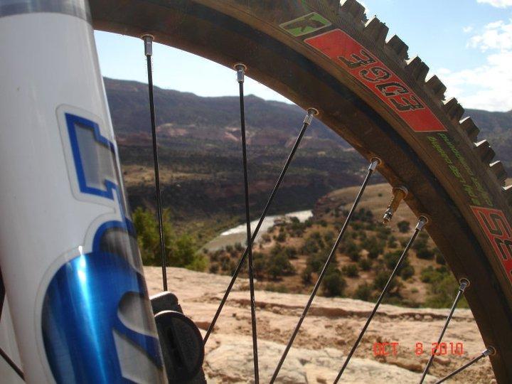 A close-up view of a mountain bike wheel with visible tire tread, set against a scenic background of mountains and a winding river. The photo captures the bike's front fork, while the landscape features greenery and layered rock formations under a clear blue sky. Mary's Loop / Horsethief Bench mountain bike trail.