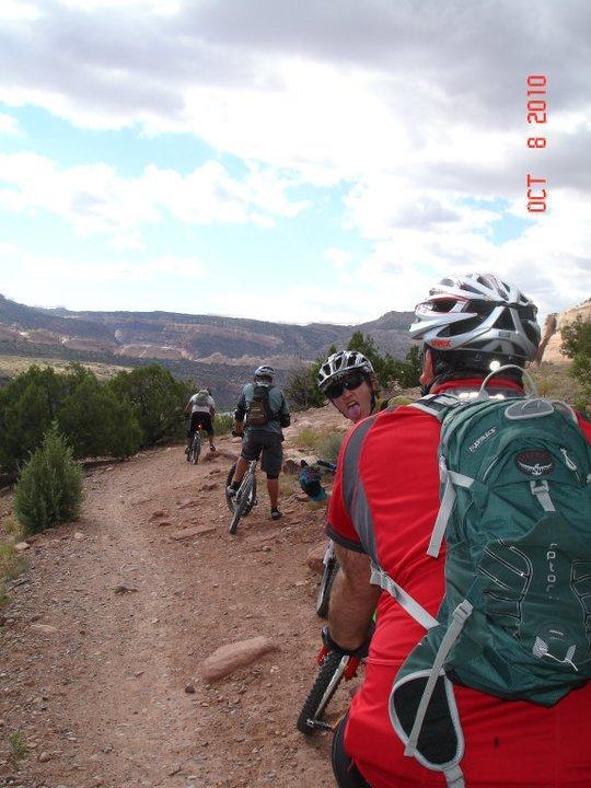 A group of mountain bikers riding along a rocky trail in a scenic outdoor landscape. One rider in the foreground, wearing a red jersey and a backpack, is playfully sticking out his tongue, while others are seen riding ahead. The background features a valley with distant mountains and partly cloudy skies. Mary's Loop / Horsethief Bench mountain bike trail.