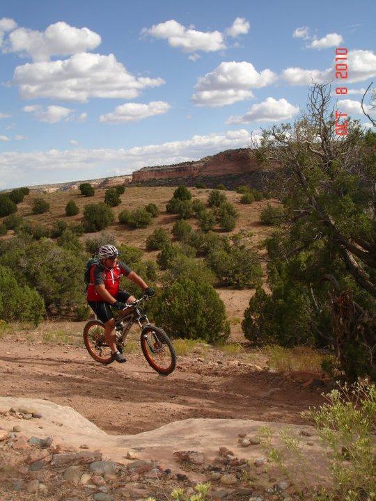 A cyclist wearing a red shirt and helmet rides a mountain bike on a dirt trail surrounded by green shrubs and trees, with a rocky landscape and blue sky with clouds in the background. Mary's Loop / Horsethief Bench mountain bike trail.
