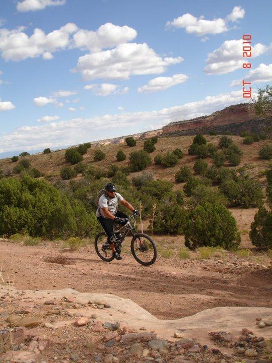 A person riding a mountain bike down a dirt trail surrounded by sparse greenery and a distant rocky landscape under a blue sky with fluffy clouds. The scene captures a sense of adventure and outdoor activity. Mary's Loop / Horsethief Bench mountain bike trail.