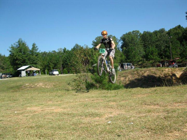 A cyclist wearing a helmet and race number jumps over a small hill on a mountain bike in a grassy area, with trees and parked vehicles in the background under a clear blue sky. Georgia International Horse Park mountain bike trail.