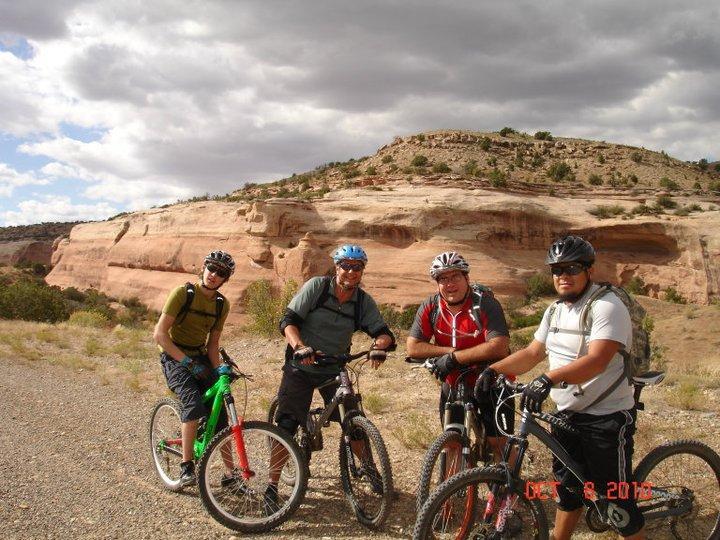 Four mountain bikers pose for a photo along a dirt trail, surrounded by scenic rock formations and cloudy skies. The group is dressed in cycling gear and stands next to their bikes, with vibrant colors and a rugged landscape in the background. Mary's Loop / Horsethief Bench mountain bike trail.