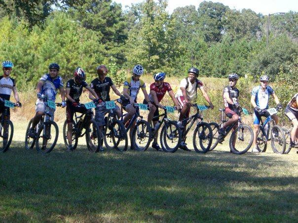 A group of eleven young cyclists lined up on mountain bikes, wearing helmets and cycling jerseys. They are positioned on a grassy area with trees in the background, ready for a race or cycling event. Each cyclist has a race number displayed on their bike. Georgia International Horse Park mountain bike trail.