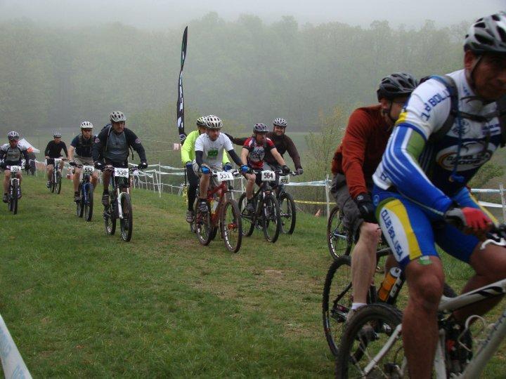 A group of mountain bikers participating in a race on a foggy day, riding along a grassy trail. Several cyclists are visible, showcasing various bike styles and racing numbers. A banner can be seen in the background, and lush trees are partially obscured by the misty atmosphere. Greenbrier State Park mountain bike trail.