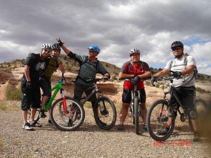 A group of five mountain bikers poses for a photo outdoors, with rocky hills and cloudy skies in the background. They are wearing helmets and biking gear, each standing beside their bicycles. One individual is playfully raising their arm, while the others smile at the camera. Mary's Loop / Horsethief Bench mountain bike trail.