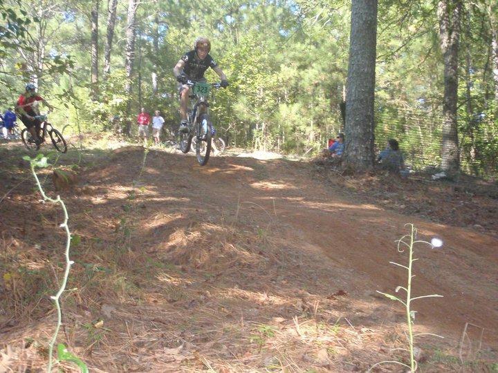 A mountain biker in a black jersey with the number 322 jumps off a dirt ramp, while another rider in a red jersey follows closely behind. The scene is set in a wooded area, with spectators visible in the background among the trees. Georgia International Horse Park mountain bike trail.