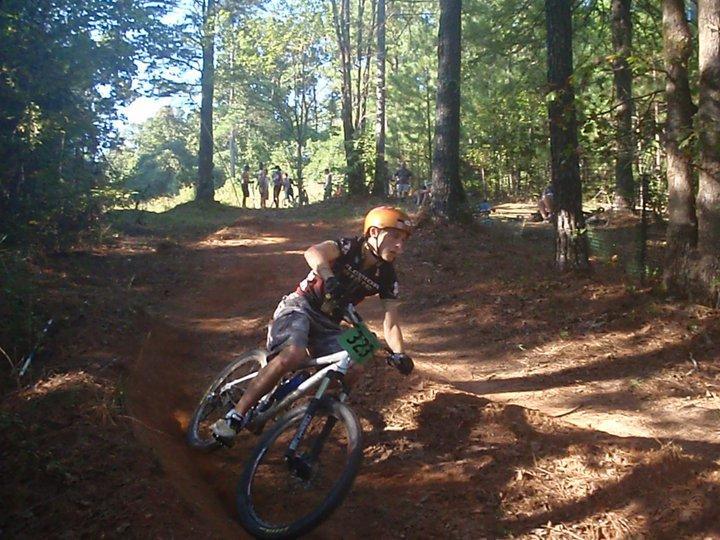 A young cyclist wearing a helmet and race number 338 leans into a turn on a dirt trail surrounded by tall trees. In the background, a few spectators can be seen enjoying the event. The scene captures the excitement of mountain biking in a wooded area on a sunny day. Georgia International Horse Park mountain bike trail.