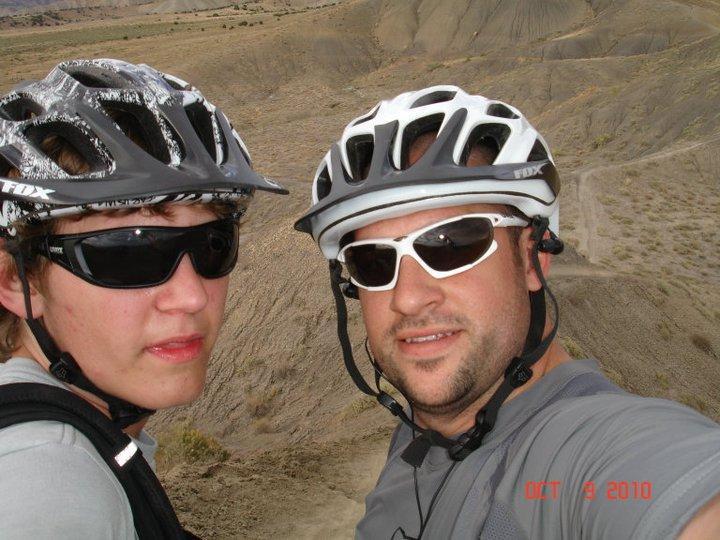 Two cyclists take a selfie while standing on a mountain trail, wearing helmets and sunglasses. The background shows a rocky landscape with hills. The photo captures a moment of adventure and camaraderie during their ride. Zippety Do Dah mountain bike trail.