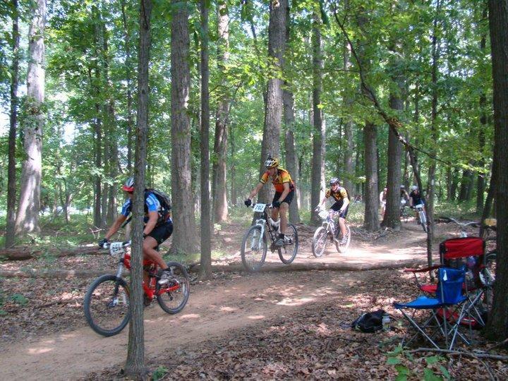 Three mountain bikers ride along a dirt trail in a wooded area, with trees surrounding them. One biker in a blue jersey and helmet is in the foreground, followed closely by another biker wearing a yellow and black jersey. A third biker trails behind, while a few spectators are seated nearby in lawn chairs. The scene captures a sunny day during an outdoor biking event. Schaeffer Farms mountain bike trail.