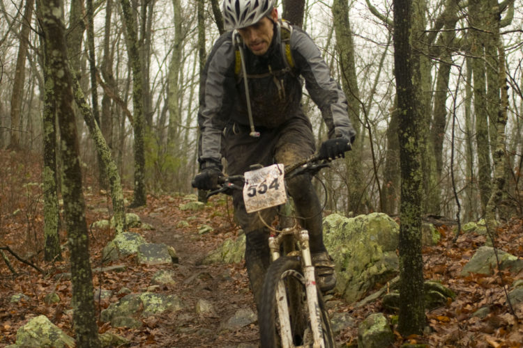A mountain biker, covered in mud, navigates a rocky, wooded trail during a rainy day. The cyclist wears a helmet and a gray jacket, with a race number visible on the bike. Surrounding trees are bare, and wet leaves cover the ground.