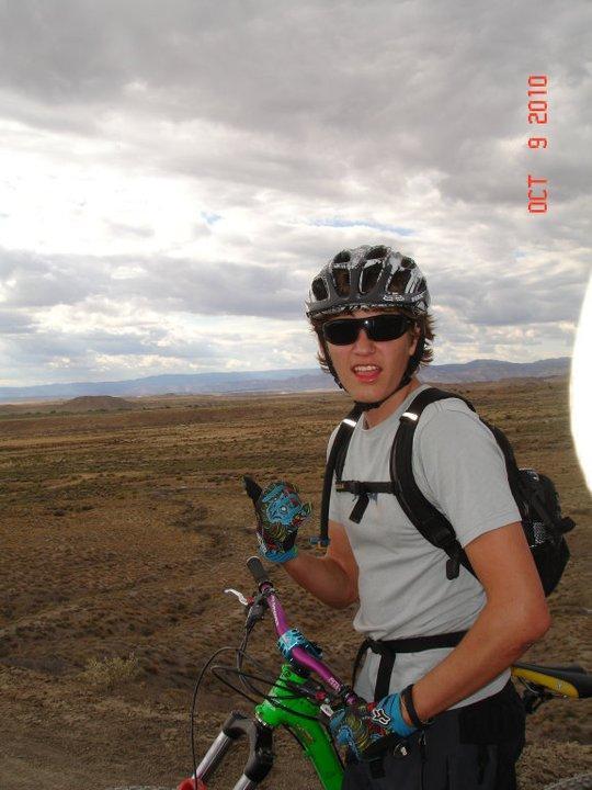 A person wearing a helmet and sunglasses poses with a mountain bike in a rugged outdoor setting. The individual is giving a thumbs-up sign while dressed in a light-colored shirt and colorful gloves. The background features expansive, dry plains under a partly cloudy sky. Zippety Do Dah mountain bike trail.