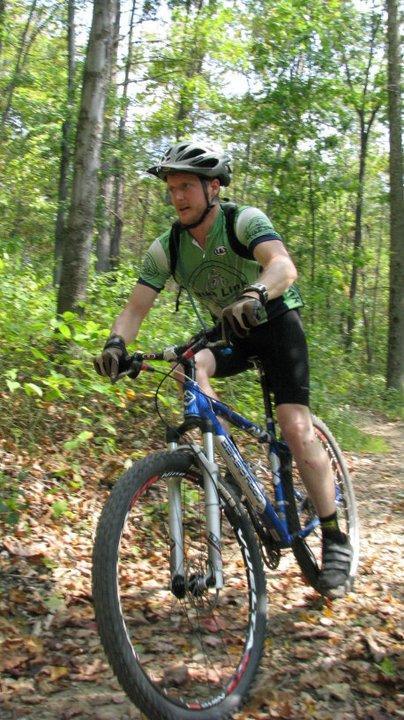A mountain biker riding on a dirt trail surrounded by green trees and foliage, wearing a helmet and a green cycling jersey, with a focused expression on his face. Allegrippis Trails mountain bike trail.