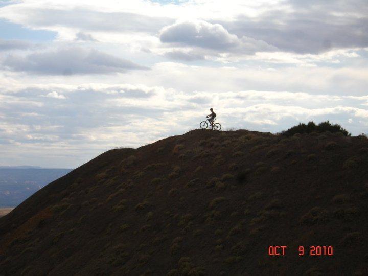 A solitary cyclist riding atop a large, rolling hillside against a backdrop of cloudy skies. The date "October 9, 2010" is displayed in the bottom right corner. Zippety Do Dah mountain bike trail.
