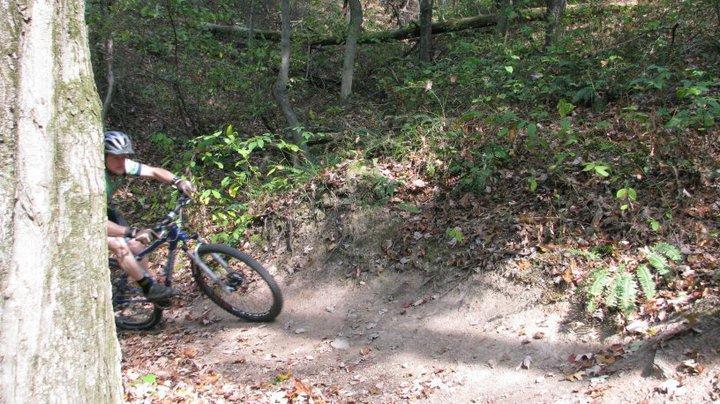 A mountain biker navigating a dirt trail surrounded by trees and autumn foliage, with a partially obscured view of the rider near a bend in the path. Allegrippis Trails mountain bike trail.