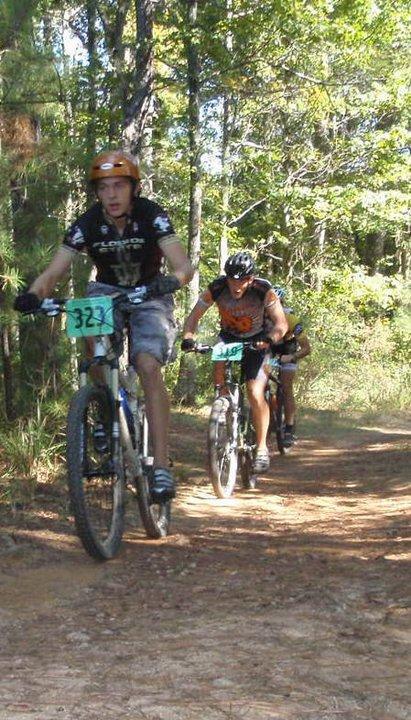 A group of three mountain bikers riding along a dirt trail, surrounded by trees. The first biker is wearing an orange helmet and a black jersey with the number 323, followed by two others in lighter gear. Dust is being kicked up from the trail as they navigate their way through the natural scenery. Georgia International Horse Park mountain bike trail.