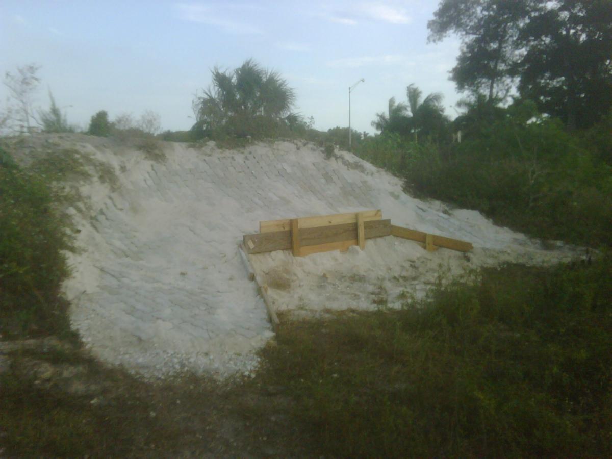 A sandy slope with wooden barriers in a natural, grassy environment, under a clear sky with scattered clouds. The area is surrounded by vegetation and palm trees. Quiet Waters Park mountain bike trail.