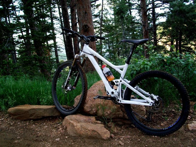 A white mountain bike leaning against a rock in a wooded area, surrounded by tall trees and greenery. The bike features thick tires and has a water bottle mounted on the frame. Chimney Gulch mountain bike trail.