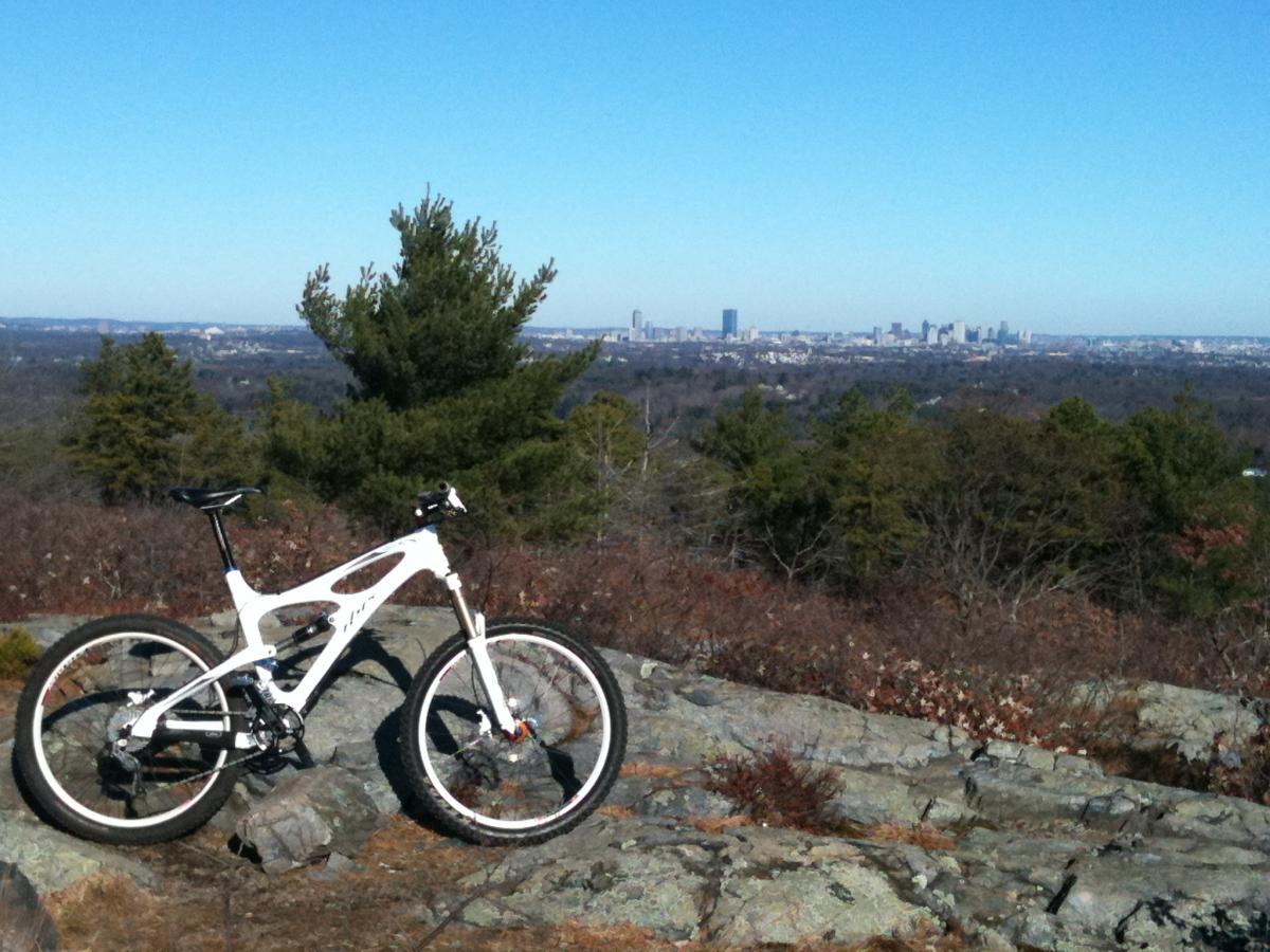 A white mountain bike rests on rocky terrain, overlooking a vast landscape with a sprawling city skyline in the background under a clear blue sky. Pine trees and rolling hills surround the scene, suggesting an outdoor adventure setting. Blue Hills mountain bike trail.
