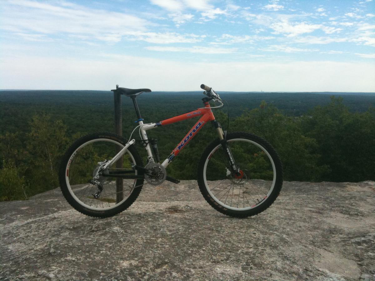 A mountain bike with an orange and white frame is positioned on a large rock outcrop, overlooking a vast landscape of lush green trees and rolling hills in the background, under a partly cloudy sky. Bradbury Mt State Park mountain bike trail.