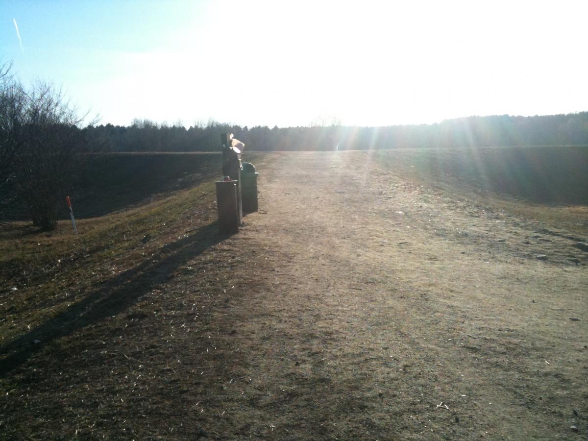 A sunlit dirt path leading towards a distant tree line, with a couple of garbage bins on the side, and a brushy area on the left. The scene is illuminated by sunlight, creating a warm atmosphere. Callahan State Park mountain bike trail.