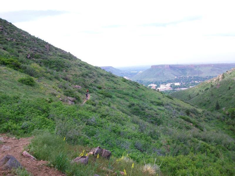 A scenic view of a lush, green hillside with a narrow dirt trail winding through it. In the distance, a small town can be seen nestled in the valley, surrounded by rolling hills under a cloudy sky. A solitary hiker, dressed in bright clothing, walks along the trail, offering a sense of scale to the expansive landscape. Chimney Gulch mountain bike trail.