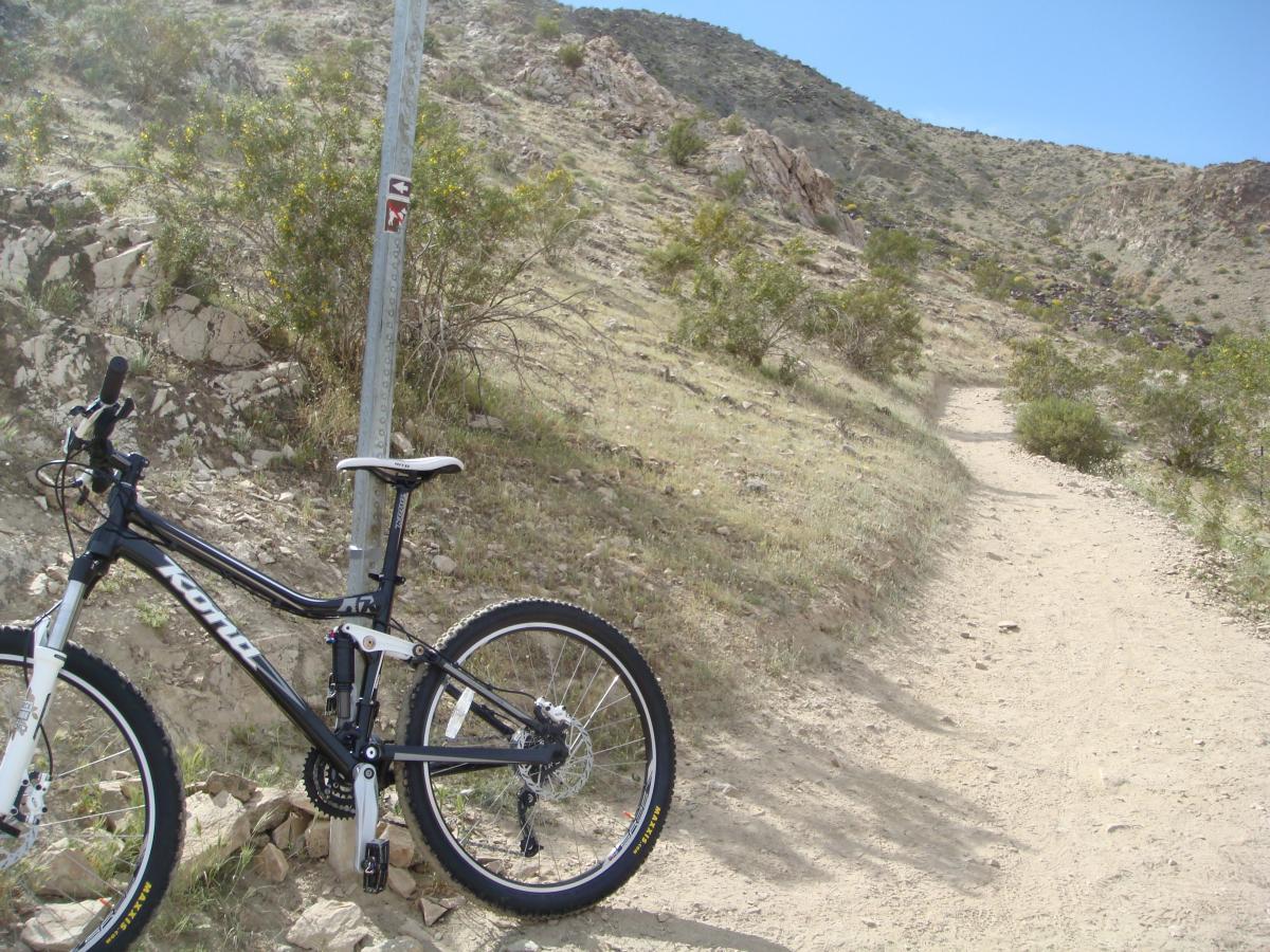 Mountain bike parked next to a trail sign on a dirt path winding through a rocky terrain and sparse vegetation under a blue sky. Bump and Grind / Hop Along mountain bike trail.