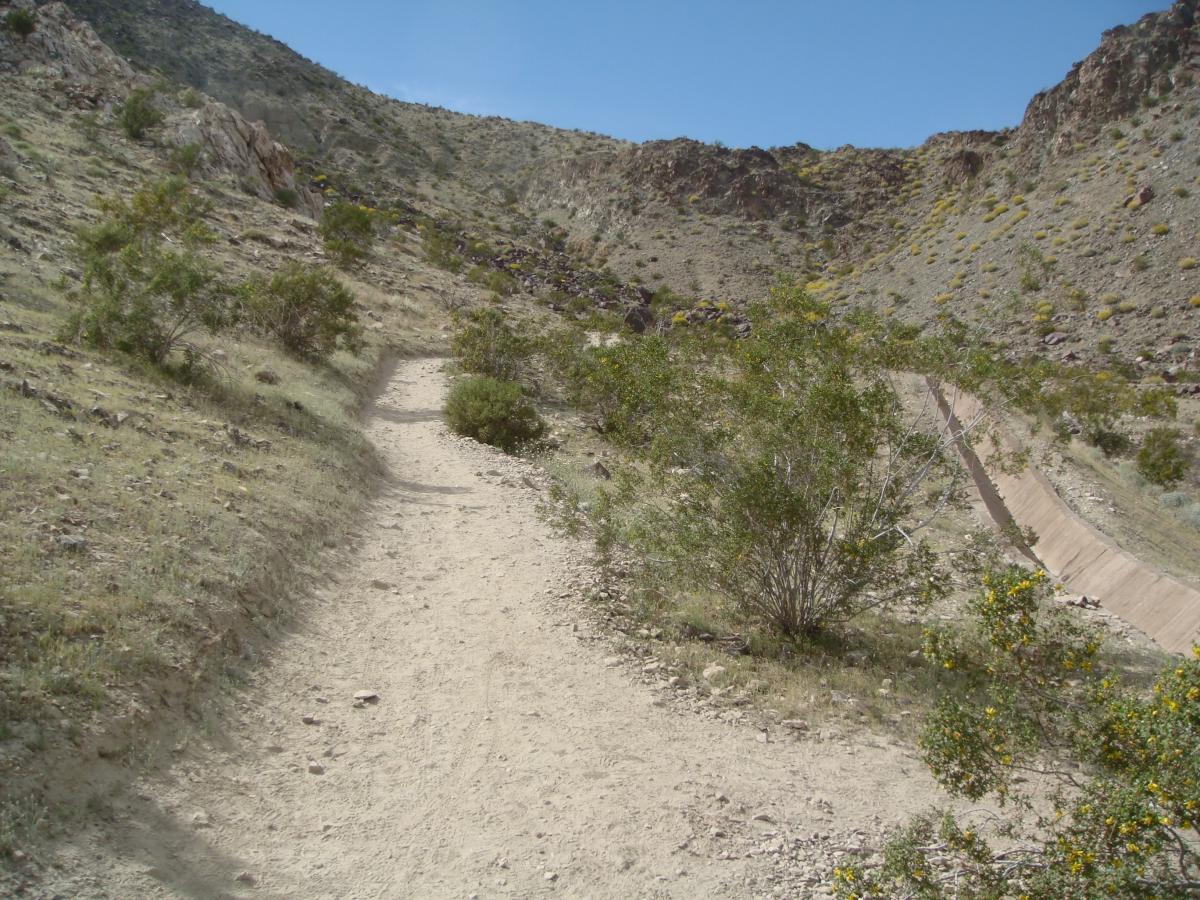 A winding dirt path leads up a hillside, surrounded by sparse vegetation and rocky terrain. In the background, a clear blue sky is visible above the mountain slopes. Bump and Grind / Hop Along mountain bike trail.