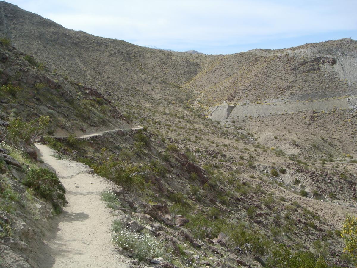 A winding dirt path trails along a rocky hillside, surrounded by sparse vegetation and rugged terrain under a partly cloudy sky. The landscape features steep, barren slopes with patches of greenery interspersed throughout. Bump and Grind / Hop Along mountain bike trail.