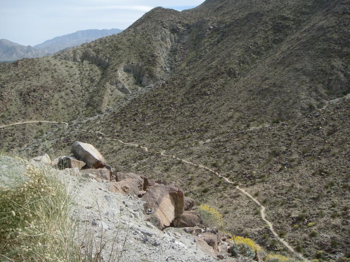 A rocky landscape with rolling hills covered in sparse vegetation, featuring a winding dirt trail that cuts through the terrain. In the foreground, large boulders and dry grass are visible, while distant mountains rise in the background under a partly cloudy sky. Bump and Grind / Hop Along mountain bike trail.