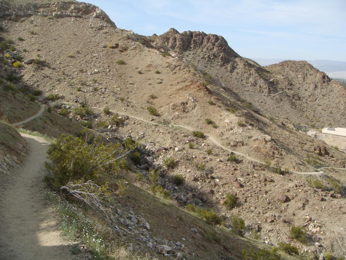 A winding dirt path meanders through a rocky, arid landscape with sparse vegetation, leading through hills and canyons under a clear blue sky. The terrain shows a mix of light brown dirt, scattered rocks, and small bushes. A person can be seen in the distance walking along the trail. Bump and Grind / Hop Along mountain bike trail.