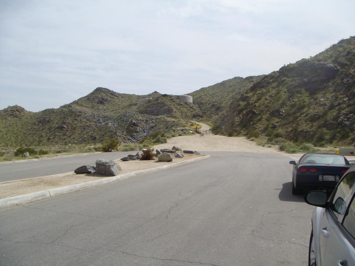 A view of a winding road leading into rocky, mountainous terrain, with sparse vegetation and a clear sky. Two parked cars are visible on the side of the road, with large rocks scattered nearby. The landscape features gentle hills and a structure in the distance at the end of the road. The Goat Trails mountain bike trail.