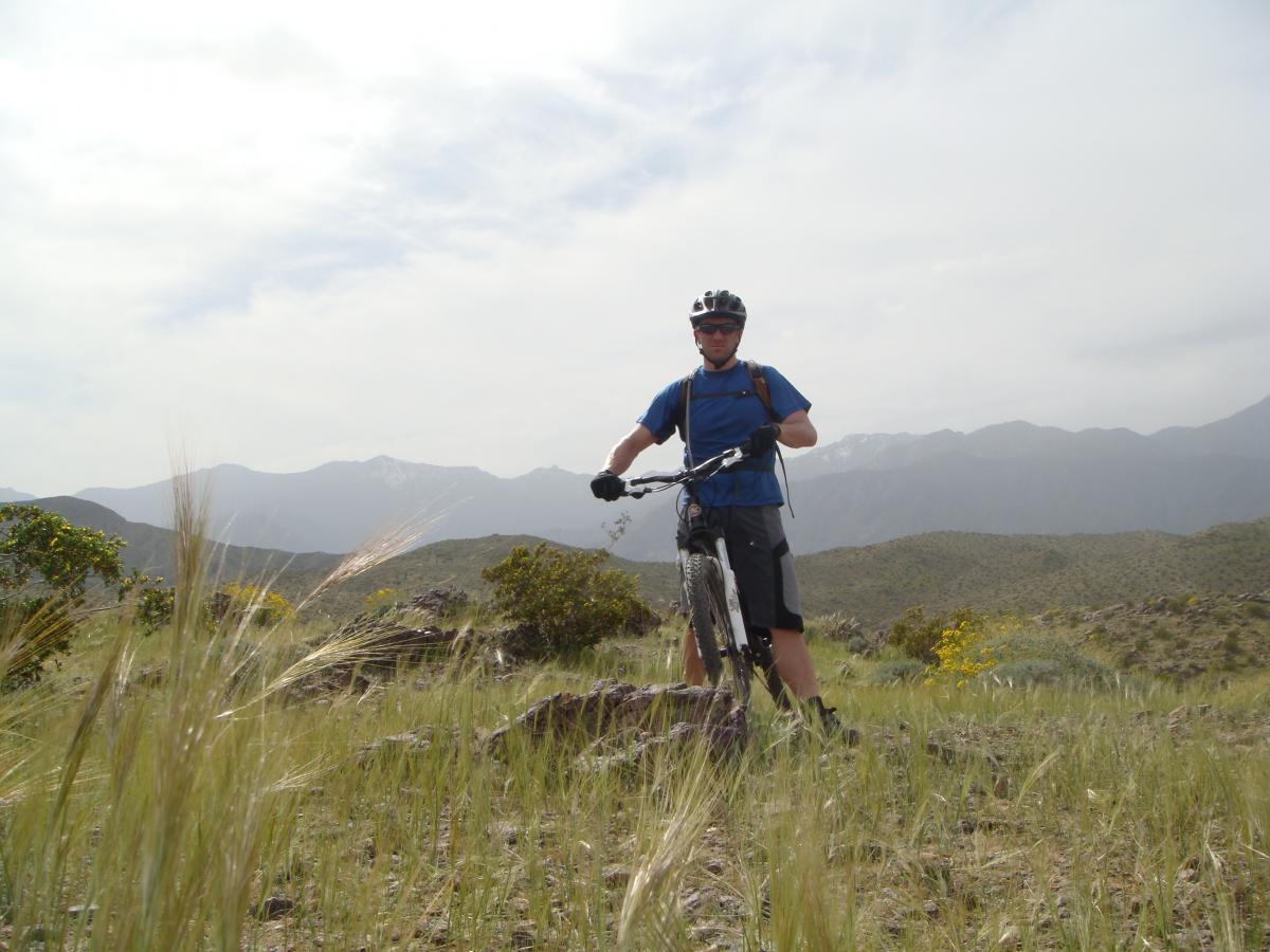 A person wearing a helmet and biking gear stands beside a mountain bike on a rocky trail, surrounded by grass and small shrubs. In the background, rolling hills and distant mountains are visible under a cloudy sky. The Goat Trails mountain bike trail.